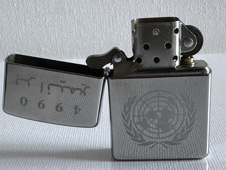 May include: A silver-colored lighter with the lid open, revealing engraved text and numbers. The body of the lighter features a detailed emblem. The lighter is a classic design, suitable for various uses.