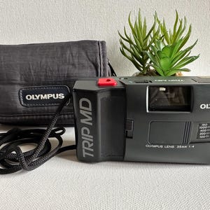 Puede incluir: Una cámara de película Olympus Trip MD negra de 35 mm con botón rojo, correa negra y funda a juego. La cámara tiene el logotipo de Olympus y Trip MD impresos. La lente es de 35 mm 1:4.