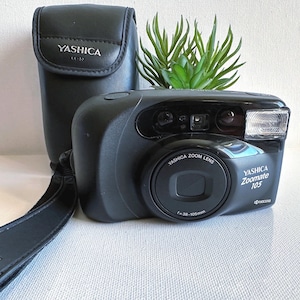 Puede incluir: Cámara negra Yashica Zoomate 105 con funda negra a juego. La cámara tiene un objetivo zoom y un flash incorporado. La funda tiene el logotipo de Yashica. Una correa negra está sujeta a la cámara.