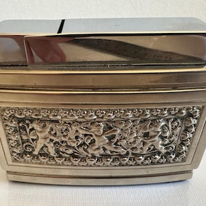 Puede incluir: Un pitillera plateada con forma rectangular y tapa con bisagras. La parte delantera de la pitillera presenta un diseño en relieve detallado de querubines y elementos florales. La tapa superior es de color plateado pulido.