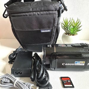 Puede incluir: Videocámara Canon LEGRIA HF R16 negra con una funda de transporte Cullmann negra, cargador, cables y una tarjeta SD SanDisk Extreme. La videocámara tiene una lente y las palabras "8.0 MEGA PIXELS" impresas.