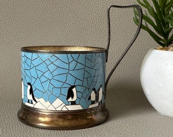 Puede incluir: Un portavasos de esmalte plateado y azul con asa. El portavasos tiene un diseño de mosaico con fondo azul y detalles blancos. El diseño presenta una fila de pingüinos en blanco y negro.