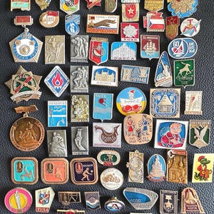 Puede incluir: Una colección de pines y insignias vintage de la era soviética, con varias formas y diseños. Los pines muestran una gama de colores, incluyendo rojo, azul, dorado y plateado, con algunos mostrando texto y símbolos. La colección está dispuesta sobre una superficie oscura.