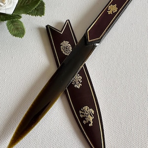 Op de afbeelding: Een briefopener met een donkerbruin handvat en een bijpassende schede. Het handvat heeft een gouden rand en een gouden embleem. De schede is donkerbruin met gouden versieringen, waaronder een leeuw en een wapen.