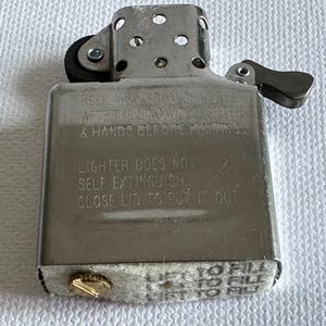 May include: A silver metal lighter with a hinged lid and a small wheel for ignition. The lighter has text engraved on the front, including safety instructions. The bottom has a gold-colored screw and a felt pad.