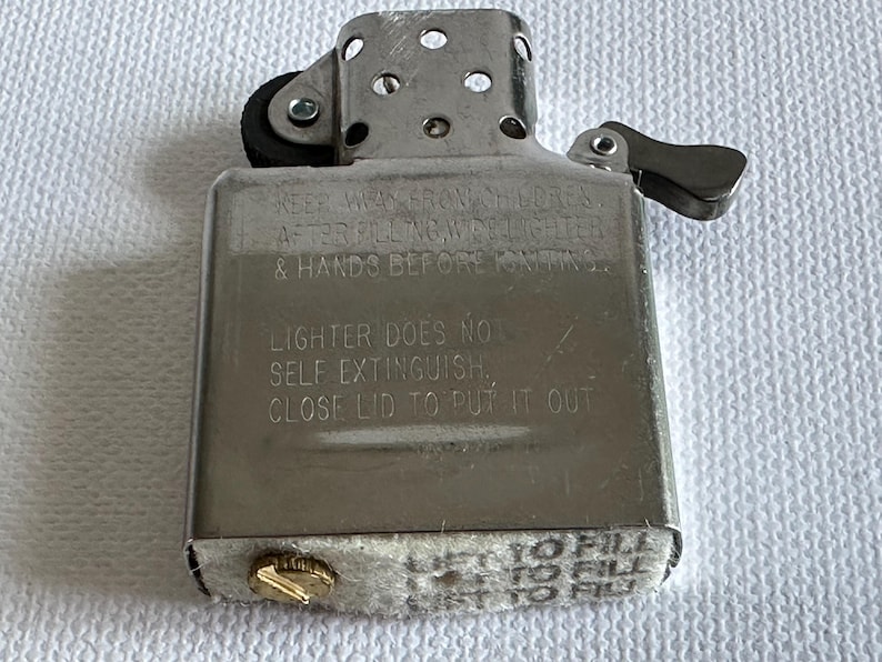 May include: A silver metal lighter with a hinged lid and a small wheel for ignition. The lighter has text engraved on the front, including safety instructions. The bottom has a gold-colored screw and a felt pad.