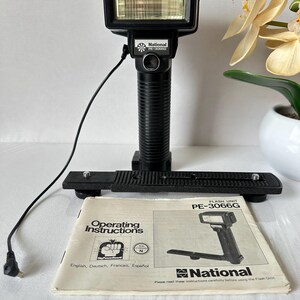 Puede incluir: Un flash National PE-3066G negro con una luz rectangular y un mango acanalado. La unidad está conectada a una base negra y a un cable de alimentación. Un manual de instrucciones está frente al flash.