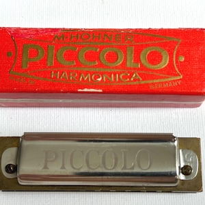 Pode incluir: Uma harmónica Piccolo de metal prateado com a palavra PICCOLO gravada na frente, acompanhada pela sua caixa de cartão vermelha original com letras douradas que dizem M. HOHNER PICCOLO HARMONICA MADE IN GERMANY.