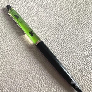 Op de afbeelding: Een zwarte en zilveren pen met een doorzichtige houder gevuld met groene vloeistof en zwarte silhouetten. De pen heeft een zwarte punt en een zilveren clip. De pen ligt op een getextureerd, lichtgrijs oppervlak.