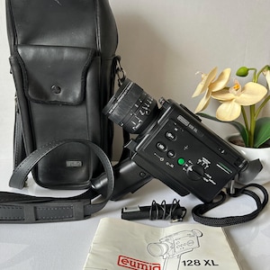 Puede incluir: Una cámara de cine Super 8 vintage Eumig 128 XL negra con una funda de transporte de cuero negro y un manual de instrucciones. La cámara tiene un objetivo zoom y una variedad de controles.