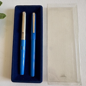 May include: Two blue fountain pens with silver accents are displayed in a dark blue velvet case. A clear plastic case is next to the pen case. The pens are a vibrant blue color.