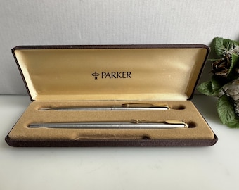 Puede incluir: Un juego de bolígrafos Parker plateados en un estuche marrón con interior dorado. El estuche está abierto, revelando dos bolígrafos con detalles dorados. La palabra "PARKER" está impresa en dorado en el interior del estuche.