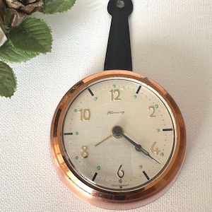 May include: A round wall clock with a copper-colored frame and a cream-colored face. The dial features gold-colored numerals for 2, 4, 6, 8, 10, and 12, with the word Blessing printed in black script. A black metal tab extends from the top.