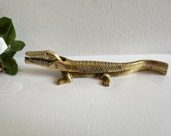 Puede incluir: Figura de caimán de latón con mandíbulas abiertas, escamas detalladas y patas pequeñas. La escultura de estilo vintage es un objeto decorativo, probablemente un cascanueces o un abridor de botellas. El caimán mide unos 25 cm de largo.