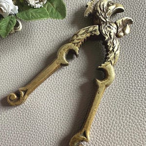 May include: A vintage brass nutcracker with an eagle head design. The nutcracker has two curved handles and a detailed eagle head at the center. The brass has an aged, antique finish. The background is a textured, light gray surface.