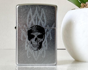 Puede incluir: Encendedor plateado con un diseño de calavera y huesos cruzados. La calavera lleva un pañuelo y está rodeada de llamas.