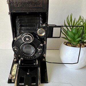 May include: A black vintage folding camera with a leather exterior and a Compur shutter. The camera has a viewfinder and a lens with a focusing ring. The camera is in good condition and has a classic look.