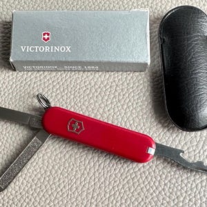 May include: A red Swiss Army knife with various tools, including a blade and file, is shown open. The knife features the Victorinox logo and is accompanied by a black leather case and a grey box. The box reads "VICTORINOX" and "THE ORIGINAL SWISS ARMY KNIFE."