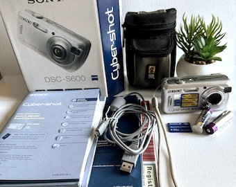 Puede incluir: Una cámara digital Sony Cyber-shot DSC-S600 plateada con su caja original, manuales y accesorios. La caja muestra la imagen de la cámara. Se incluye una funda, un cable USB y pilas.