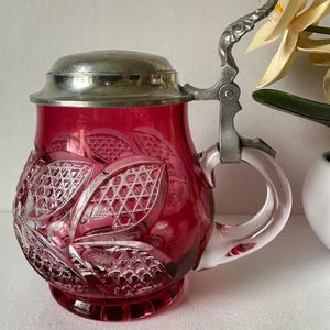 Op de afbeelding: Een robijnrode glazen bierpul met een decoratief geslepen glazen patroon. De pul heeft een zilveren metalen deksel en handvat. Het handvat is van helder glas. De pul is ongeveer 15 cm hoog.