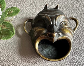 Puede incluir: Un cenicero de latón vintage con forma de cara de diablo con cuernos y boca abierta. El cenicero tiene una pátina oscura con detalles dorados y dos pequeñas asas.