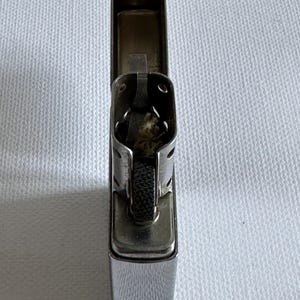 May include: A chrome-plated lighter with the top open, revealing the internal components. The lighter has a rectangular shape with rounded corners and a textured wheel for sparking. The lighter is sitting on a white surface.