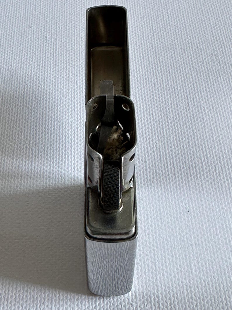 May include: A chrome-plated lighter with the top open, revealing the internal components. The lighter has a rectangular shape with rounded corners and a textured wheel for sparking. The lighter is sitting on a white surface.