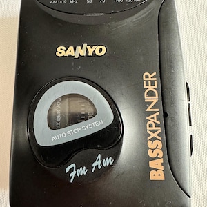 Può includere: Lettore di cassette radio portatile nero Sanyo Bassxpander. Il dispositivo ha una manopola di sintonizzazione, controllo del volume e uno sportello per cassette. Il testo "SANYO" è in lettere dorate e "BASSXPANDER" è arancione.