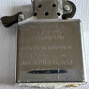 May include: A silver Zippo lighter with the text "FOR BEST RESULTS USE ZIPPO FLINTS AND FLUID" and "ZIPPO MFG. CO. BRADFORD, PA." The lighter is marked "08" and "MADE IN ZIPPO U.S.A."