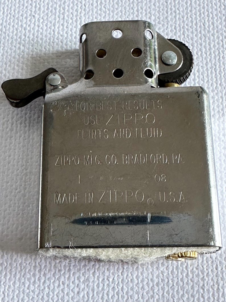 May include: A silver Zippo lighter with the text "FOR BEST RESULTS USE ZIPPO FLINTS AND FLUID" and "ZIPPO MFG. CO. BRADFORD, PA." The lighter is marked "08" and "MADE IN ZIPPO U.S.A."