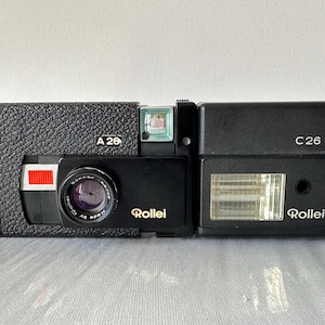 May include: A black and silver Rollei A26 camera with a built-in flash unit. The camera has a black leatherette covering and a silver lens. The flash unit is labeled "C26" and has a silver reflector.