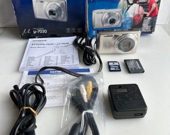 Puede incluir: Una cámara digital Olympus μ-7030 plateada con una lente negra y una correa negra. La cámara está en su caja original con un manual y accesorios. La caja es azul y plateada con el logotipo de Olympus y el nombre del modelo de la cámara.