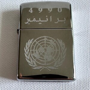 May include: A silver-colored lighter with the United Nations emblem etched on the front. The top of the lighter has the numbers "4990" and Arabic script. The lighter is rectangular and has a shiny finish.