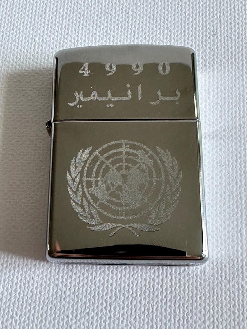 May include: A silver-colored lighter with the United Nations emblem etched on the front. The top of the lighter has the numbers "4990" and Arabic script. The lighter is rectangular and has a shiny finish.