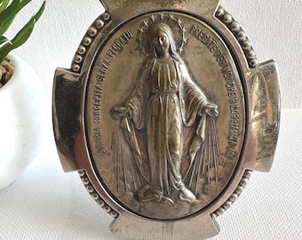 May include: A silver-toned religious pendant with an oval center featuring a detailed relief of a figure with outstretched arms. The pendant has a cross-shaped design with text around the oval. The overall design is ornate and detailed.