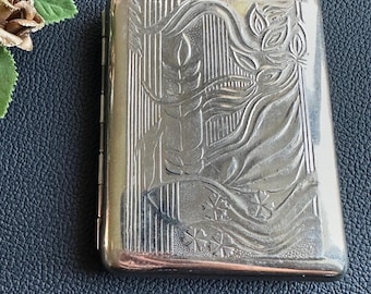 Puede incluir: Un pitillera plateada con un diseño Art Déco. La pitillera presenta una imagen en relieve de una mujer con el pelo suelto, una vela y detalles florales. La pitillera es rectangular con bordes redondeados y una tapa con bisagras.