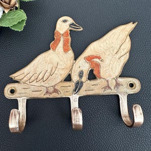 May include: A brass wall hook rack featuring two ducks with orange bows. The ducks are positioned on a horizontal bar that includes three curved hooks for hanging items. The metal has a polished finish with engraved details on the duck figures.