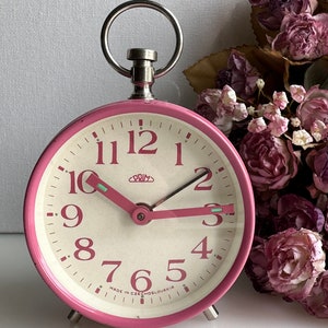 May include: A pink vintage alarm clock with a white face and black numbers. The clock has a silver handle and says "PRIM" and "MADE IN CZECHOSLOVAKIA" on the face.