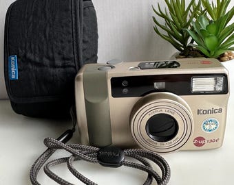 Puede incluir: Una cámara compacta Konica Z-up 130E de color dorado con una correa negra y un cordón gris. La cámara tiene un objetivo Konica con un rango de zoom de 38-130 mm.
