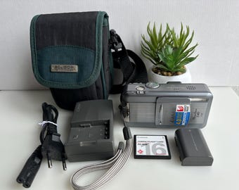 Puede incluir: Un kit de cámara digital con una cámara Canon PowerShot S40 gris, una funda Soligor negra y verde, un cargador, una batería y una tarjeta CompactFlash de 16 MB. También hay una pequeña planta en maceta.