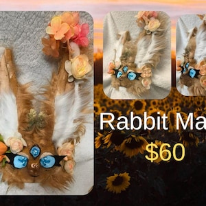May include: A brown and white rabbit mask with large blue eyes, adorned with pink and peach-colored flowers. The mask has long, upright ears and is made of faux fur. The text "Rabbit Mask" is displayed below the mask.