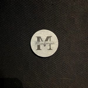 Könnte beinhalten: Eine runde, silberfarbene Münze mit dem Namen "Mackenzie" und dem Buchstaben "M" in kursiver Schrift eingraviert. Die Münze befindet sich vor einem schwarzen, strukturierten Hintergrund.