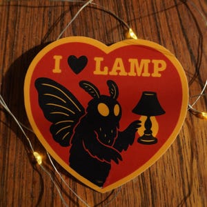 Peut inclure: Un autocollant en forme de cœur avec un fond rouge et une bordure jaune. L'autocollant représente une créature noire ressemblant à un papillon de nuit tenant une lampe, avec le texte "I ♥ LAMP" en jaune. L'autocollant est sur une surface en bois.