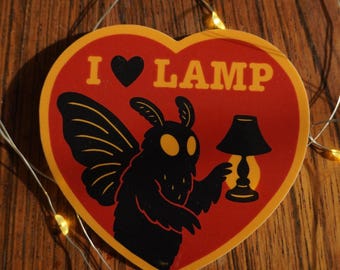 May include: A heart-shaped sticker with a red background and a yellow border. The sticker features a black moth-like creature holding a lamp, with the text "I ♥ LAMP" in yellow. The sticker is on a wooden surface.