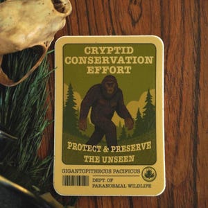 May include: A rectangular sticker with a green border and a brown background. The sticker features a dark brown illustration of a Bigfoot-like creature walking in front of green trees. Text on the sticker reads "CRYPTID CONSERVATION EFFORT" and "PROTECT & PRESERVE THE UNSEEN."