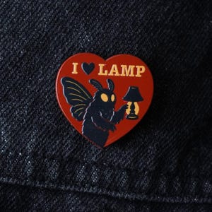 May include: A heart-shaped enamel pin with the words "I ♥ LAMP" in gold lettering. The pin features a black moth holding a lamp on a red background. The pin is set against a dark blue denim background.