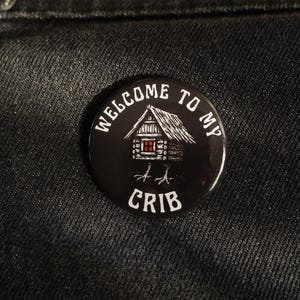 May include: A black button with the text "WELCOME TO MY CRIB" in white. The button features a cabin illustration with chicken feet and a red door. The button is round and has a glossy finish.