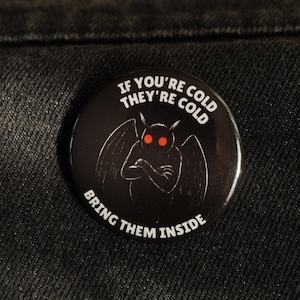 Könnte beinhalten: Runder, schwarzer Button mit dem Text "IF YOU'RE COLD THEY'RE COLD BRING THEM INSIDE" und einer Grafik einer geflügelten Kreatur mit roten Augen. Der Button hat ein glänzendes Finish.