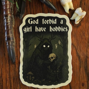 May include: A sticker with the text "God forbid a girl have hobbies" in a black and white design. The sticker features a dark forest scene with a creature holding a skull. Additional items include teeth and a decorative claw.