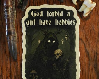 May include: A sticker with the text "God forbid a girl have hobbies" in a black and white design. The sticker features a dark forest scene with a creature holding a skull. Additional items include teeth and a decorative claw.